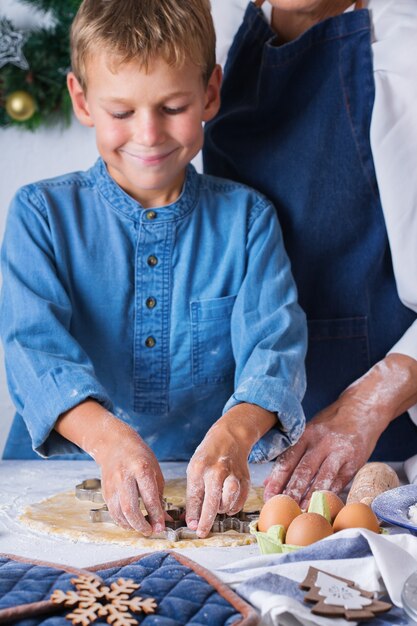 Happy senior mature woman, grandmother and young boy, grandson cooking, kneading dough, baking pie, cake,  cookies. Family time in the cozy kitchen. Seasonal winter Christmas activity at home.