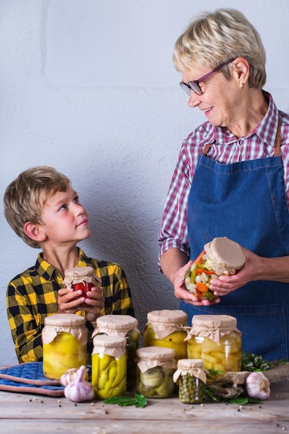 Happy senior mature woman, grandmother and young boy, grandson holding in hands jars with homemade preserved and fermented food, pickled and marinated vegetables. Harvest preservation, family time