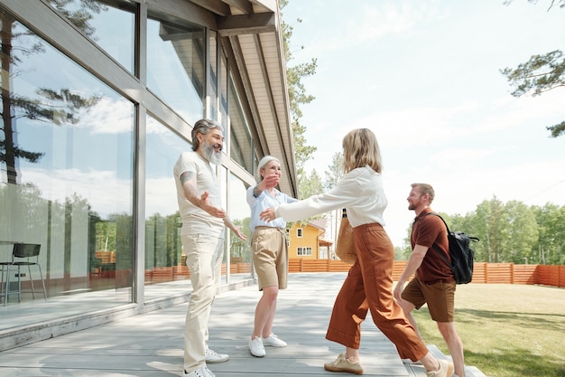 Happy senior parents outstretching arms for hugs while greeting adult son and daughter in cottage house