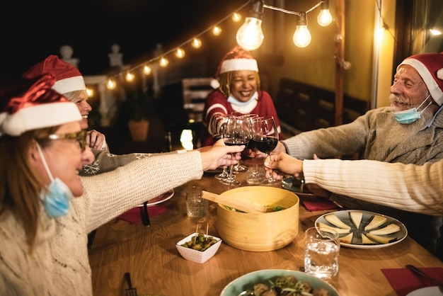 Happy senior people cheering with wine during Christmas dinner with safety masks under chins - Focus on glasses