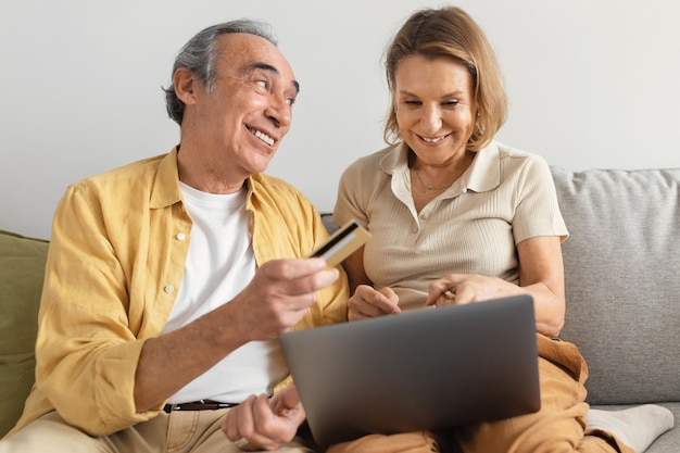 Счастливые пожилые супруги делают покупки в Интернете на ноутбуке, мужчина дает кредитную карту своей жене, сидящей дома на диване