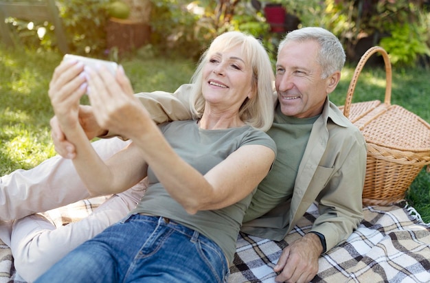 Happy senior spouses video calling on smartphone while having picnic and resting in garden at warm