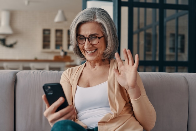 Happy senior woman in casual clothing looking at her smart phone and waving while sitting on the sofa at home