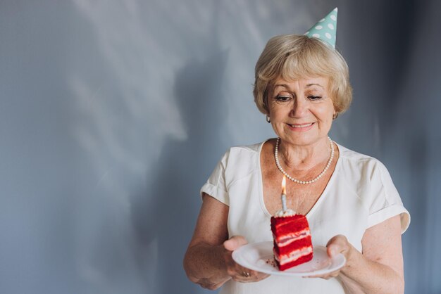 Happy senior woman celebrating her birthday wearing a party hat holding a cake with a candle on a gray background