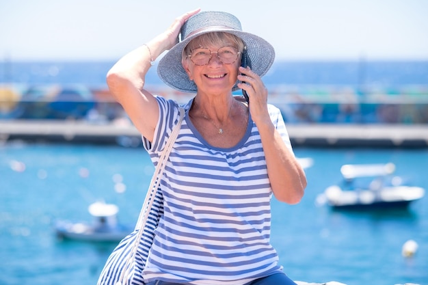 Happy senior woman dressed in blue sitting at the sea harbour holding her hat lest it fly away Mature attractive lady enjoying relaxation and vacation while using mobile phone