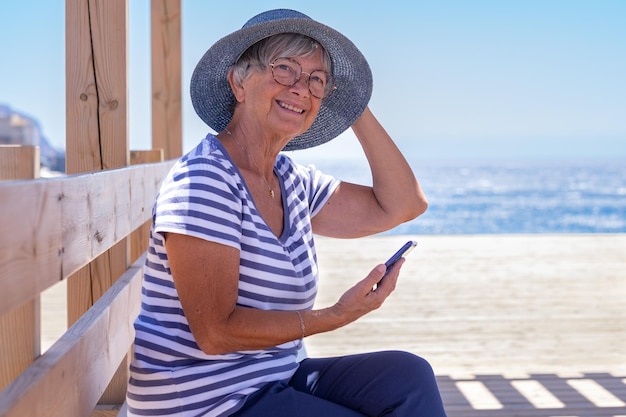 Счастливая пожилая женщина, одетая в синее, сидит у моря, держа шляпу, чтобы она не улетела Зрелая дама в очках наслаждается отдыхом и отдыхом, используя мобильный телефон