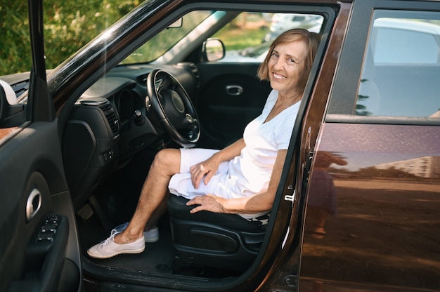 楽しんでいるカメラを見て笑って新しい茶色の車に座って運転して幸せな年配の女性