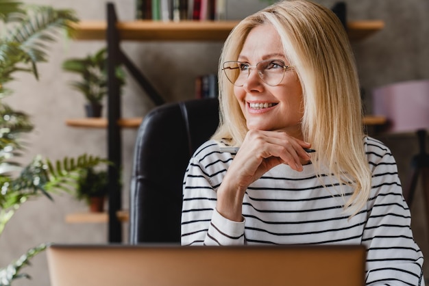 Happy senior woman holding pen daydreaming looking away sitting front of workplace