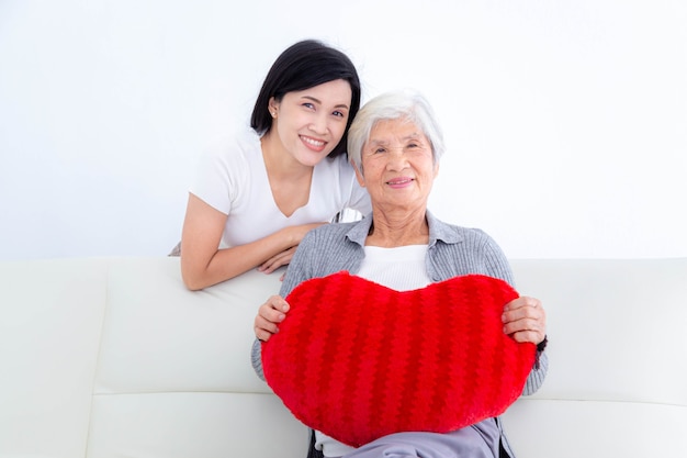 Felice donna anziana che tiene un cuscino cuore rosso con sua figlia seduta sul divano. buona festa della mamma. concetto di famiglia felice