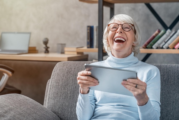 Счастливая пожилая женщина смотрит и смеется над своим цифровым планшетом на диване