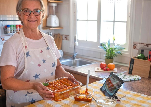 Happy senior woman shows a pan with homemade stuffed cannelloni ready for the oven Ingredients around her