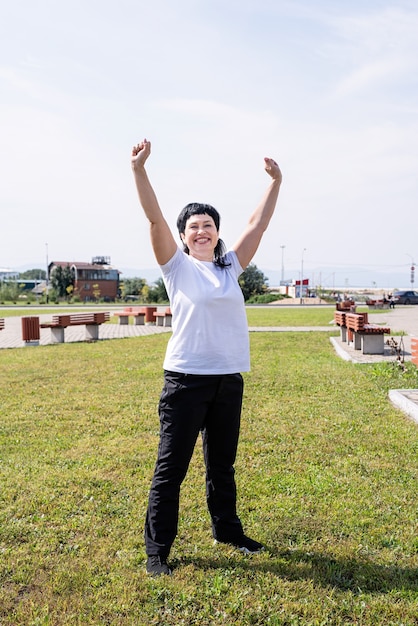 Happy senior woman in sports clothes exercising in the park standing with arms up