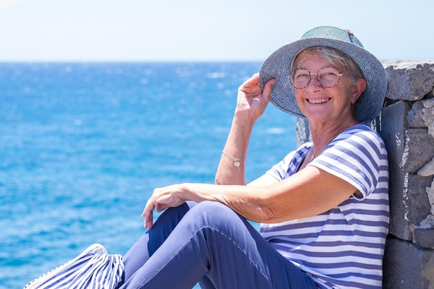 Happy senior woman in striped tshirt sitting by the sea holding her hat lest it fly away Mature lady dressed in blue enjoying relaxation and vacation looking at camera