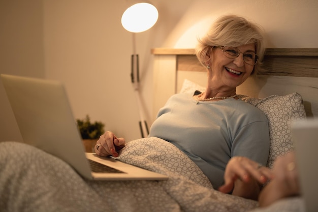 Happy senior woman surfing the net on laptop while communicating with her husband in bedroom at night