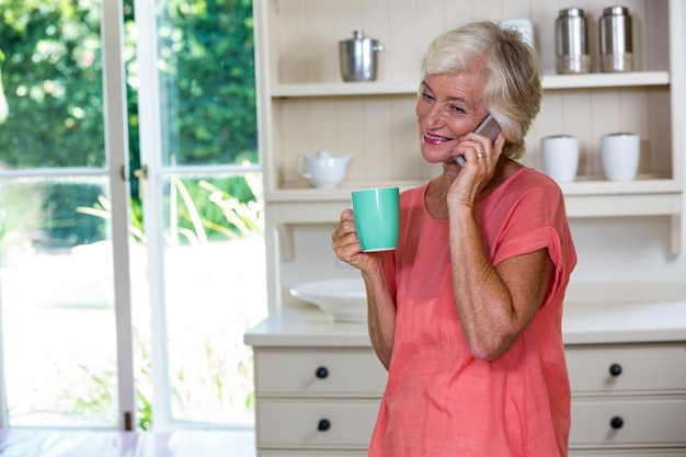 Happy senior woman talking on phone while having coffee in kitchen