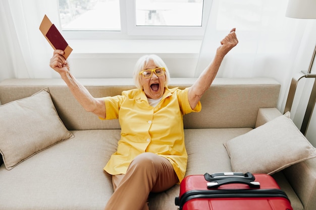 Счастливая пожилая женщина с паспортом и проездным билетом упаковала красный чемодан в отпуск и здравоохранение Улыбающаяся пожилая женщина радостно сидела на диване перед поездкой и радостно подняла руки вверх