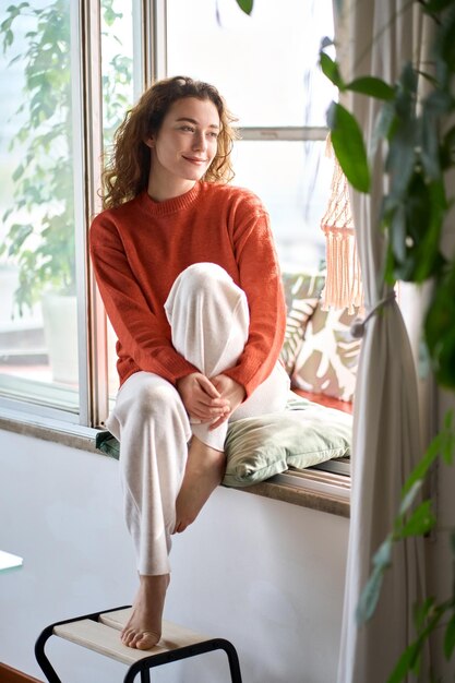 Happy serene young woman sitting relaxing at home looking through window