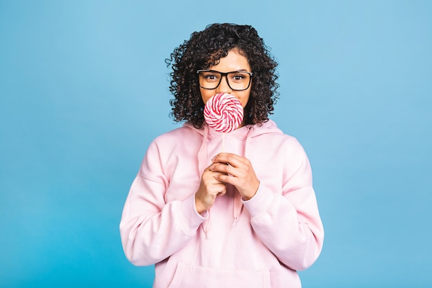 Photo happy sexy american afro girl eating lollipop. beauty glamour model woman holding sweet colorful lollipop candy, isolated on blue background. sweets.