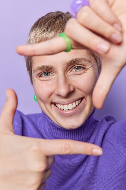 Happy short haired young woman takes photo with frame gesture\
looks through fingers as if photographing with camera smiles\
toothily wears purple turtleneck poses indoor searches\
perspective