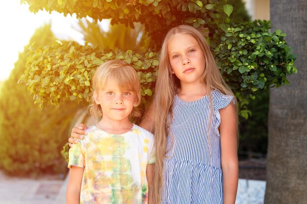 Happy sibling kids travelers boy brother of five years old and girl sister eight years old friends hugging on green plants background and look into camera travel on nature in summer vacation flare