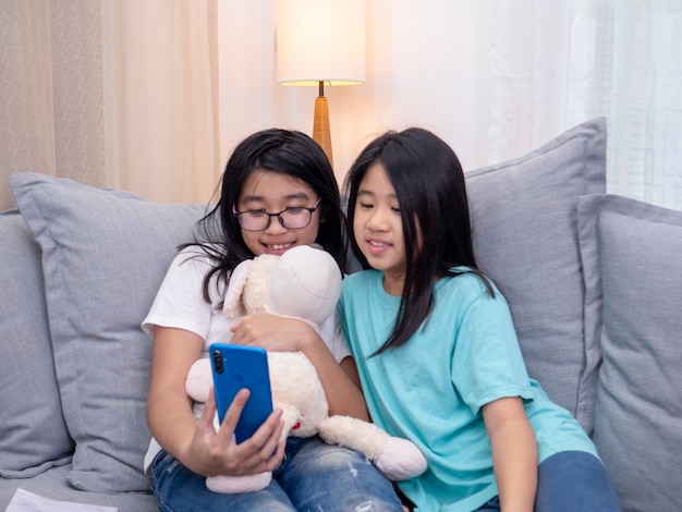 Happy siblings children sitting on sofa in living room speak with parent on mobile phone together, smiling elder girl showing cute little sister video call by smartphone speak with family.