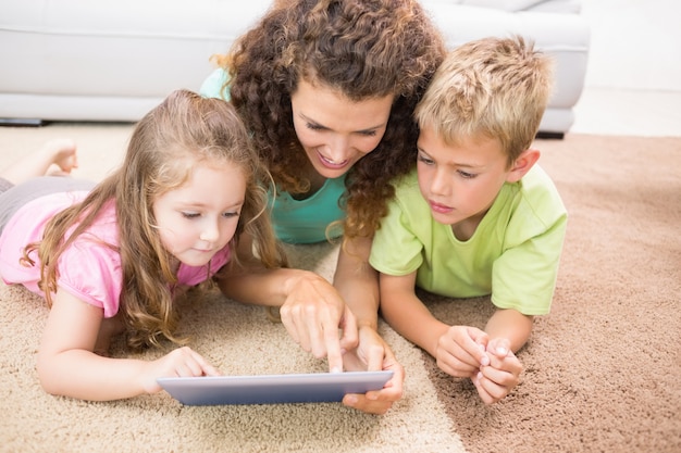 Photo happy siblings lying on the rug using tablet with their mother