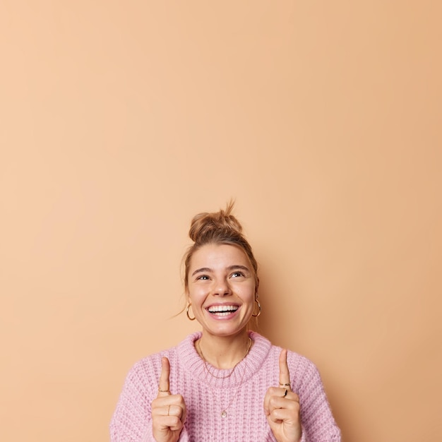 Felice e sincera giovane donna europea sorride ampiamente ai punti sopra con il dito indice indossa un maglione casual lavorato a maglia mostra un fantastico logo pubblicitario o un banner discout isolato su sfondo beige.