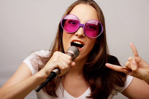 Счастливая поющая девушка. Красота женщины нося белую футболку и большие солнцезащитные очки с микрофоном на белом фоне. ipster styl