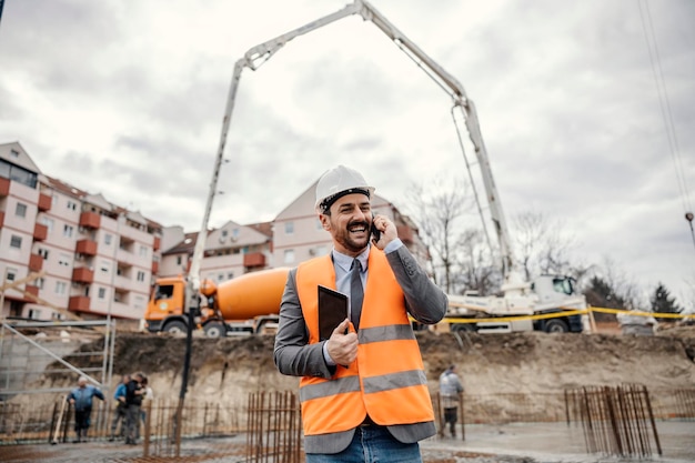 A happy site manager is holding tablet and talking on the phone