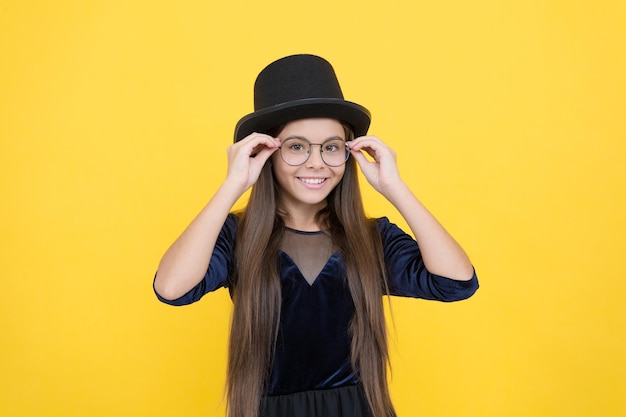 Happy small baby wear black hat on long brunette hair fixing fashion glasses with party look yellow background sight