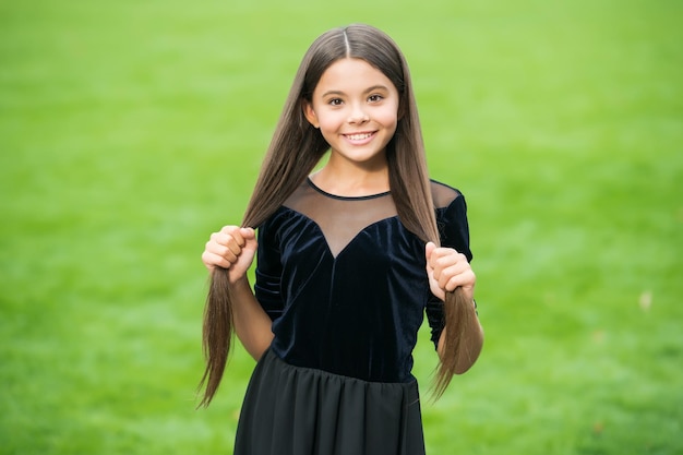 ファッションドレスの緑の草の日当たりの良い夏の屋外サロンで長いブルネットの髪を保持している美しさの表情の笑顔で幸せな小さな女の子