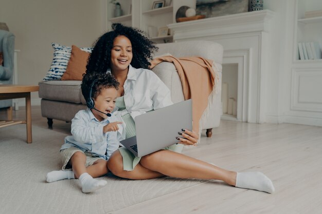 Счастливый маленький ребенок мальчик с помощью гарнитуры во время разговора онлайн на ноутбуке, сидя на полу с мамой