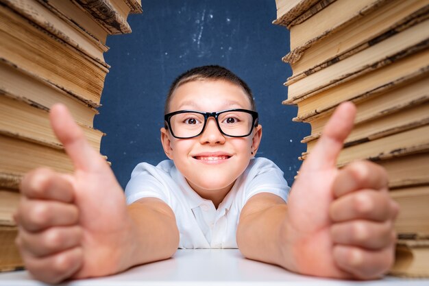 Felice ragazzo intelligente con gli occhiali seduto tra due pile di libri sorridenti e mostrando i pollici in su.