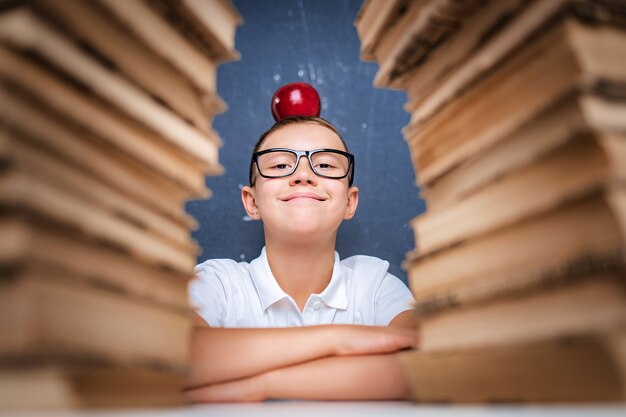 Happy smart boy in glasses sitting between two piles of books with red apple on head and look at camera smiling.