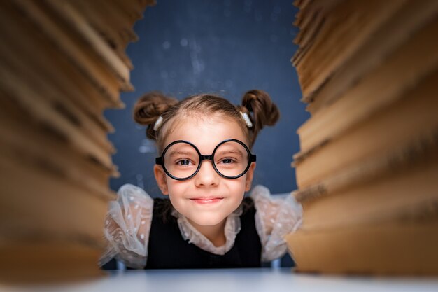 Happy smart girl in rounded glasses sitting between two piles of books and look at camera smiling.