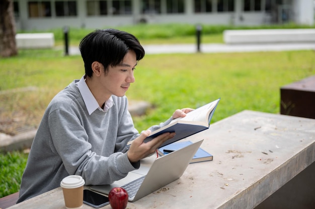 A happy and smart young Asian college man is focusing on reading a book in the campus park