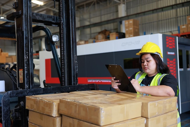 Happy smile Asian obese female warehouse worker in safety vest and hardhat helmet at warehouse factory industrial product in cardboard boxes on shelves Plus size female inspecting products working