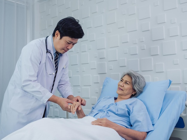 밝은 파란색 드레스를 입은 아름다운 아시아 노부인 환자가 침대에 누워 있는 동안 하얀 양복을 입은 남성 의사가 병실에서 손을 잡고 정맥 주사액을 주는 행복한 미소