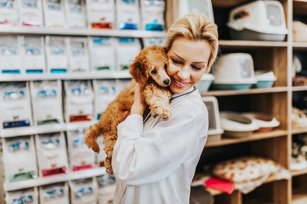 Happy and smiled middle age veterinarian woman standing in pet\
shop and holding cute miniature red poodle.