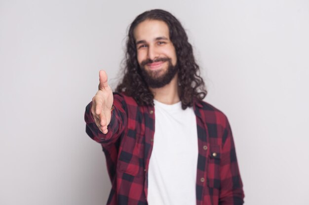 Happy smiley handsome man with beard and black long curly hair in casual style, checkered red shirt standing giving handshake and greeting with smile. indoor studio shot, isolated on grey background.