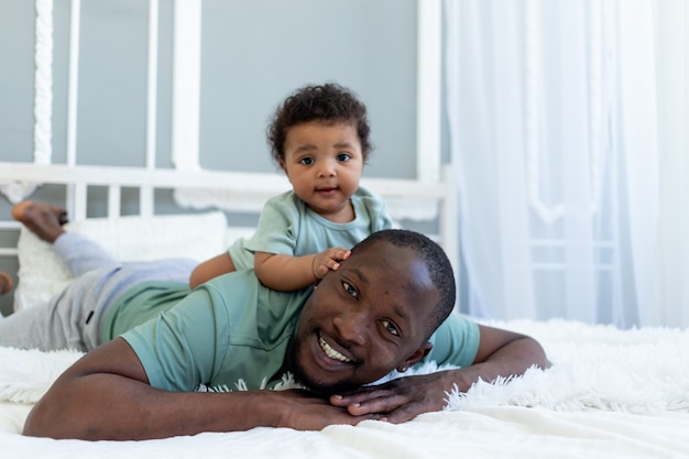 Happy smiling African american dad with baby son on bed at home cuddle, happy family