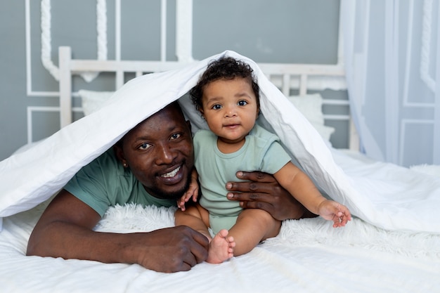 Happy smiling African american dad with baby son on the bed at home cuddling under the blanket, happy family