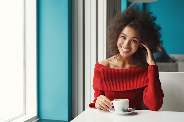 Happy smiling african-american girl at cafe table. Young dreamy woman enjoying hot coffee in pleasant atmosphere. Leisure and urban lifestyle concept.