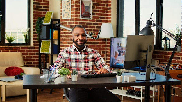 Happy smiling african american man working from home, sitting at desk in stylish cozy apartment with creative eclectic hygge interior design. Cheerful upbeat teleworker remotely doing job, close up