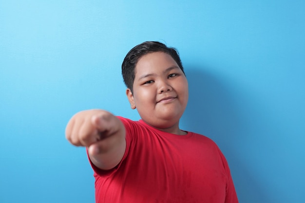 Happy smiling asian boy looking at camera and pointing boy wearing red shirt against blue background