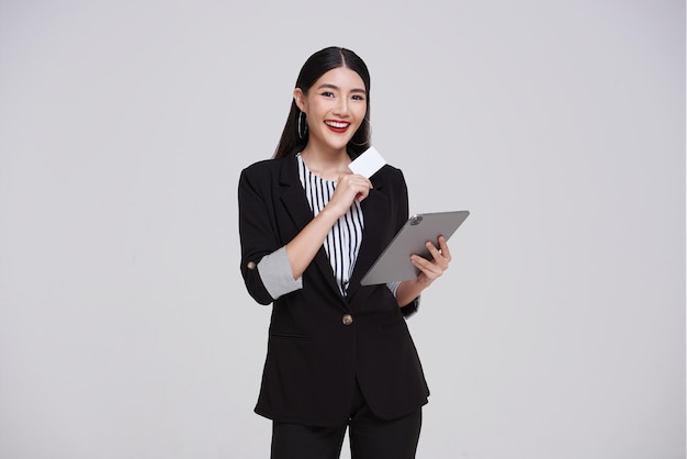 Happy smiling Asian business woman in suit using tablet and showing presenting credit card