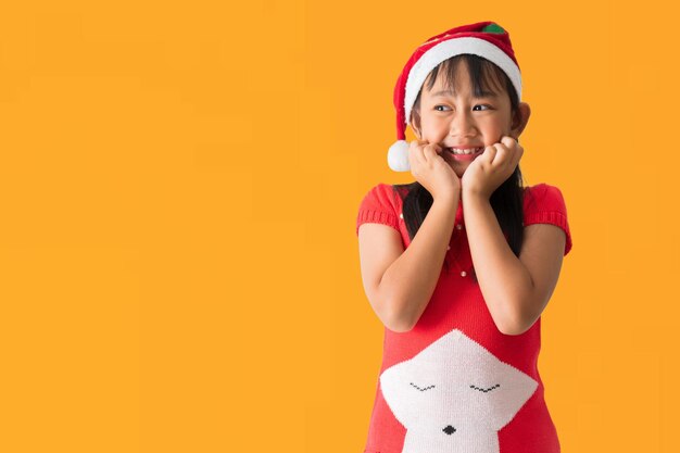 은 크리스마스 의상을 입고 산타클로스 모자를 입은 행복한 미소 짓는 아시아 어린이 소녀가 노란색 배경에 고립되어 서서 놀랐습니다.