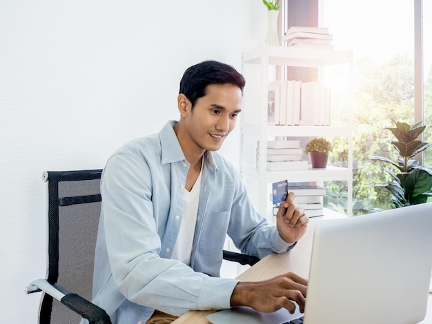 Happy smiling Asian man in denim shirt shopping online paying with a credit card in his hand with a laptop computer for his own small business on a white home office background table