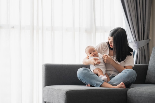 自宅のソファで娘の赤ちゃんと一緒に楽しい時間を過ごしている幸せな笑顔のアジア人の母親