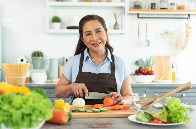 La donna più anziana asiatica sorridente felice in grembiule taglia le verdure mentre cucina l'insalata in cucina